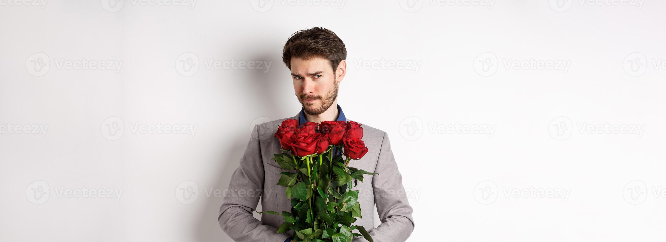 Indecisive bearded man in suit, holding flowers roses and looking at camera unsure, going on romantic date, standing over white background photo