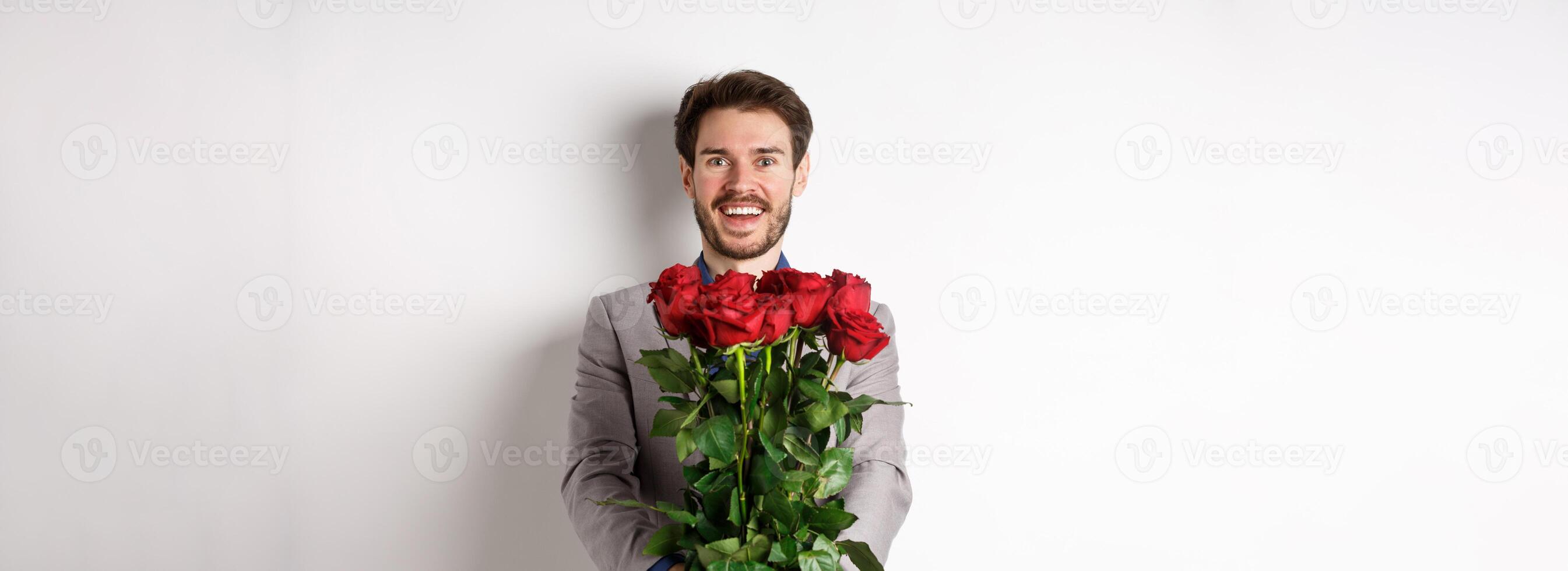 Handsome man in love wishing happy valentines day, giving bouquet of flowers on romantic date, smiling at camera, wearing suit over white background photo