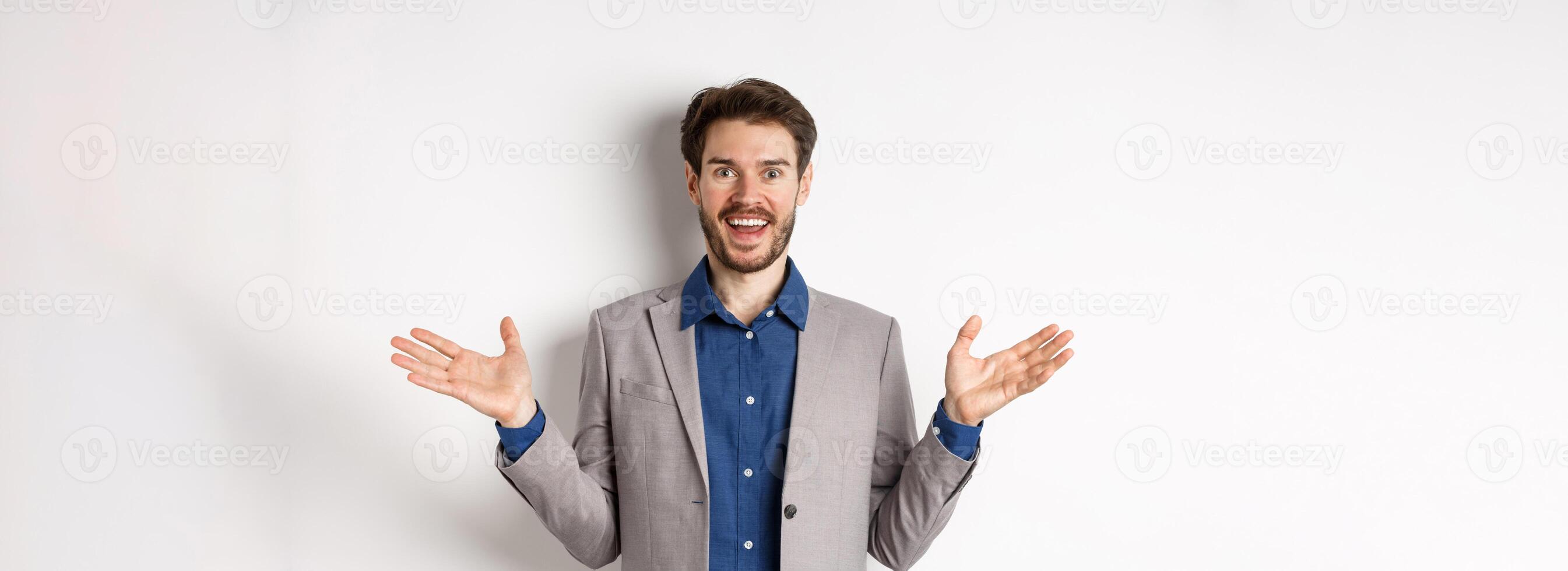 Excited male ceo manager in suit looking happy and amused, spread hands sideways to welcome or congratulate you, standing on white background photo