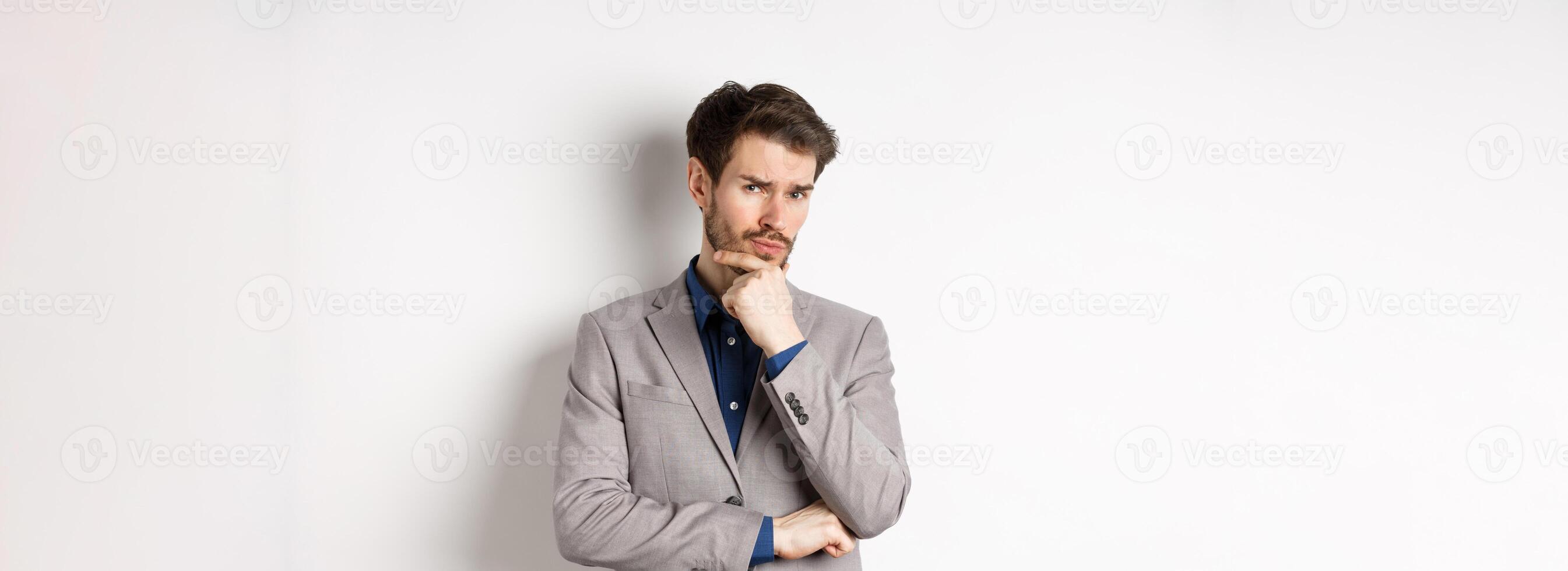 Hesitant young businessman in suit looking skeptical at camera, thinking and making decision, standing against white background photo