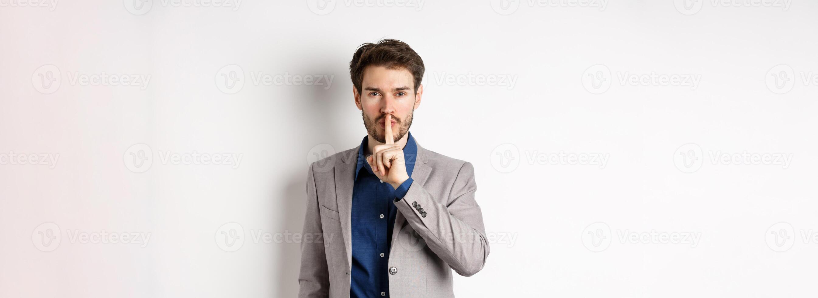 Handsome macho man in suit shushing at camera, press finger to lips, ask keep voice down, tell secret, standing against white background photo