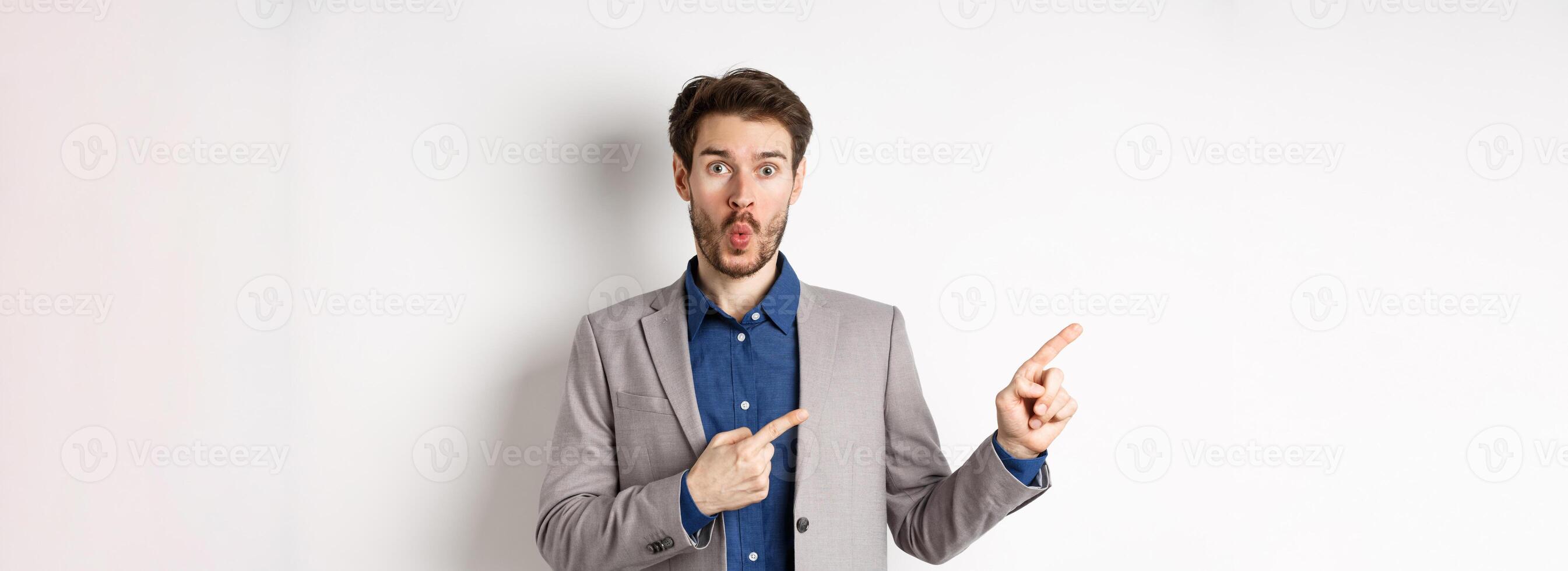 Excited handsome man in suit pointing fingers left at banner and saying wow, look here gesture, showing advertisement, standing on white background photo