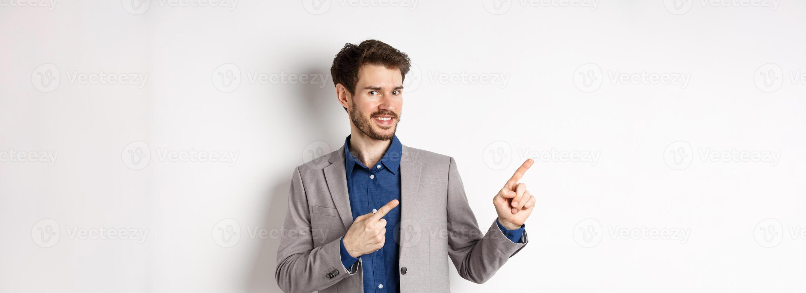 Handsome bearded businessman in grey suit pointing fingers left at logo, inviting check out advertisement, standing on white background photo
