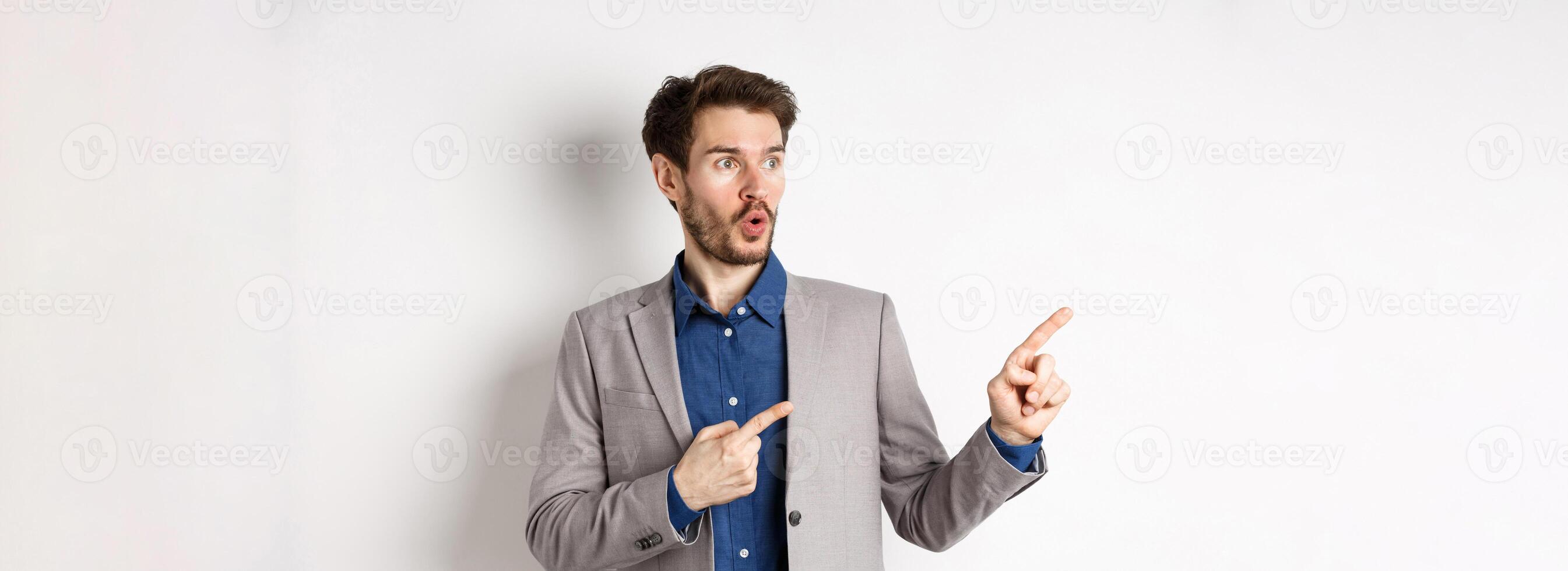 Wow look there. Excited male ceo manage in suit pointing fingers left, looking at logo impressed, showing advertisement, standing on white background photo