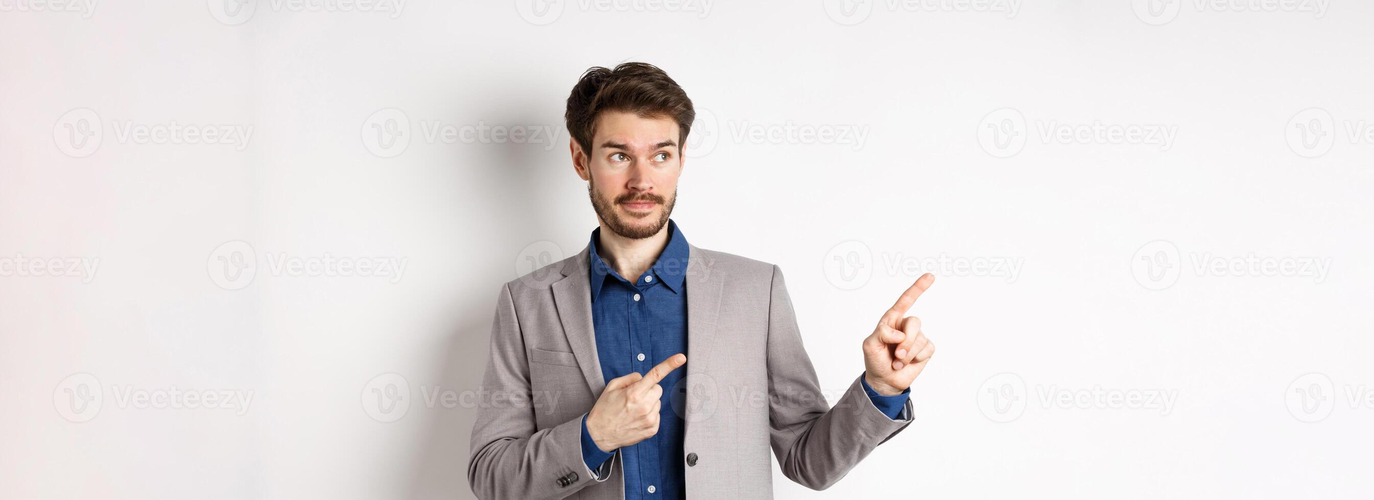 Doubtful male entrepreneur in suit looking and pointing left with hesitant face, standing unsure against white background photo