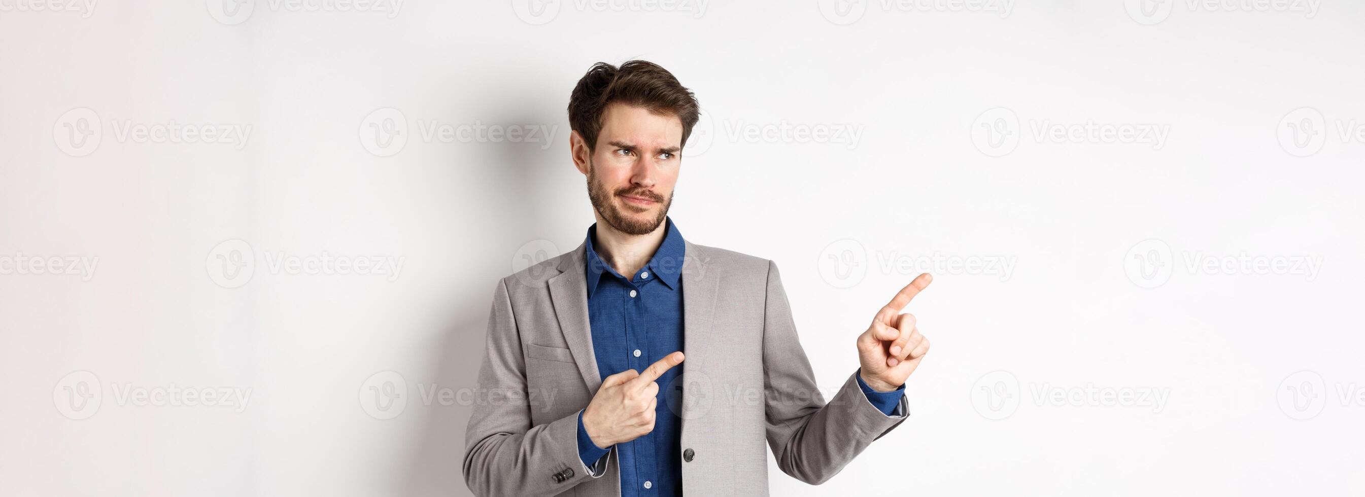 Displeased businessman in suit pointing and looking left with skeptical smirk, dislike banner, disapprove promo offer, standing on white background photo