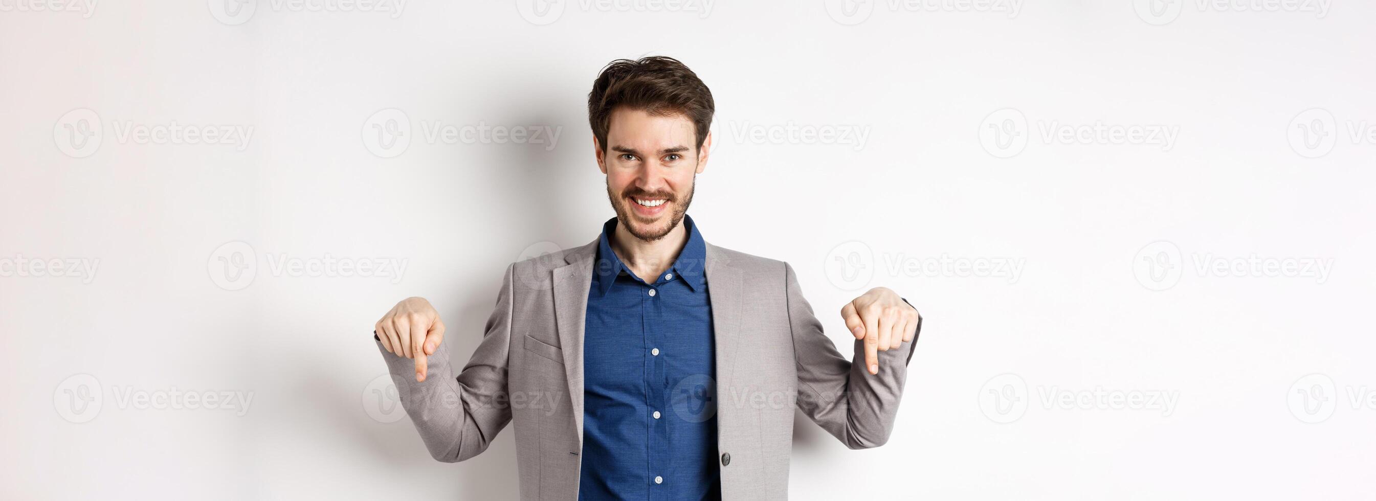 Smiling handsome man in business suit pointing fingers down, showing good deal, look here gesture, standing on white background photo
