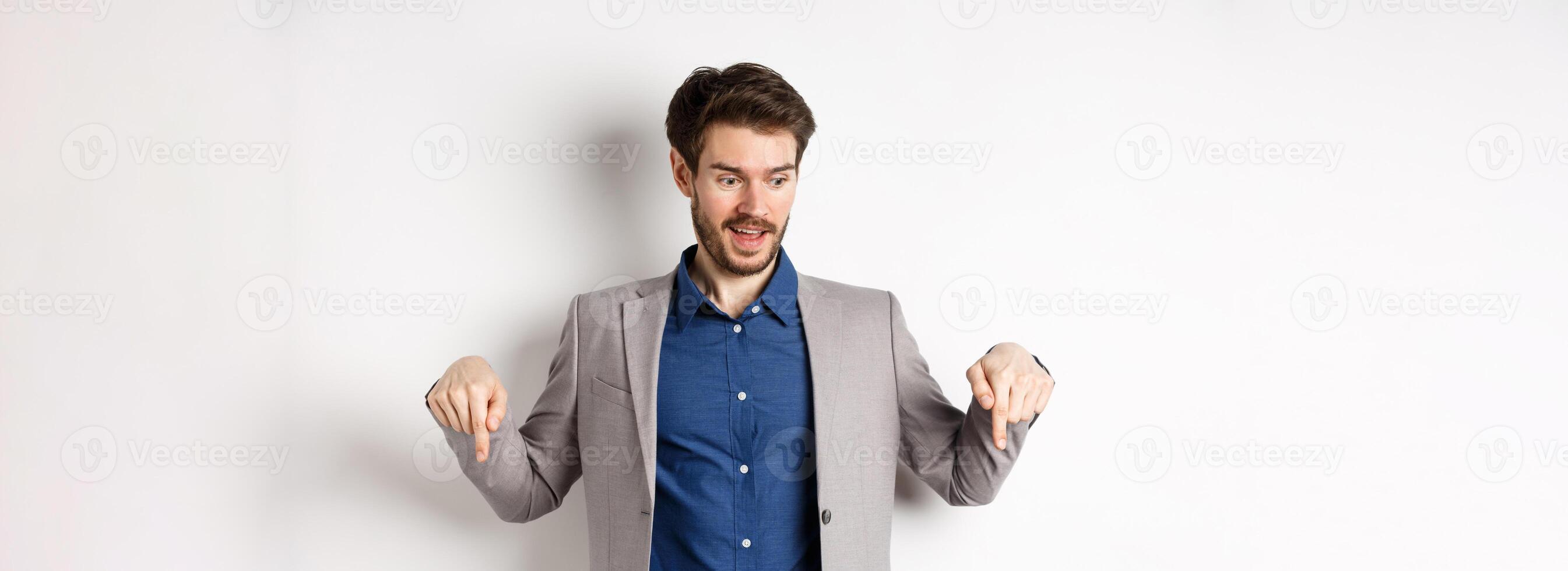 Excited guy in suit smiling and pointing fingers down, looking at special deal with amused face, standing on white background photo
