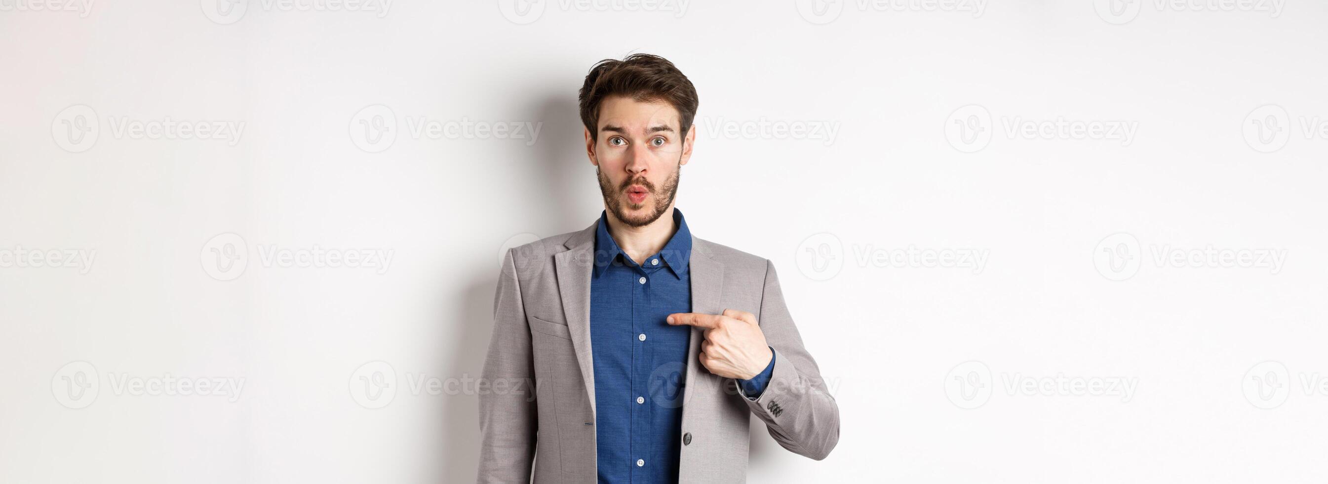 Who me. Surprised man in suit pointing at himself with disbelief, looking confused at camera, standing on white background photo