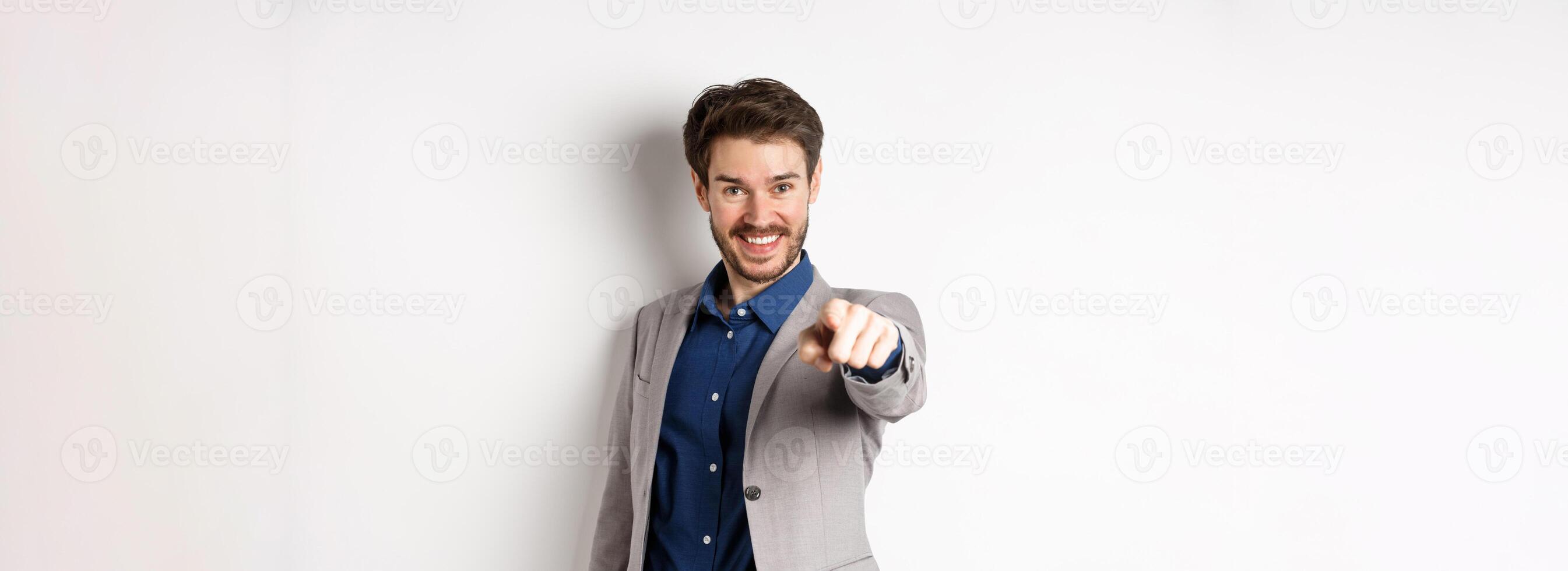 Its you. Smiling handsome manager in suit pointing at camera, choosing person and looking pleased, standing on white background photo