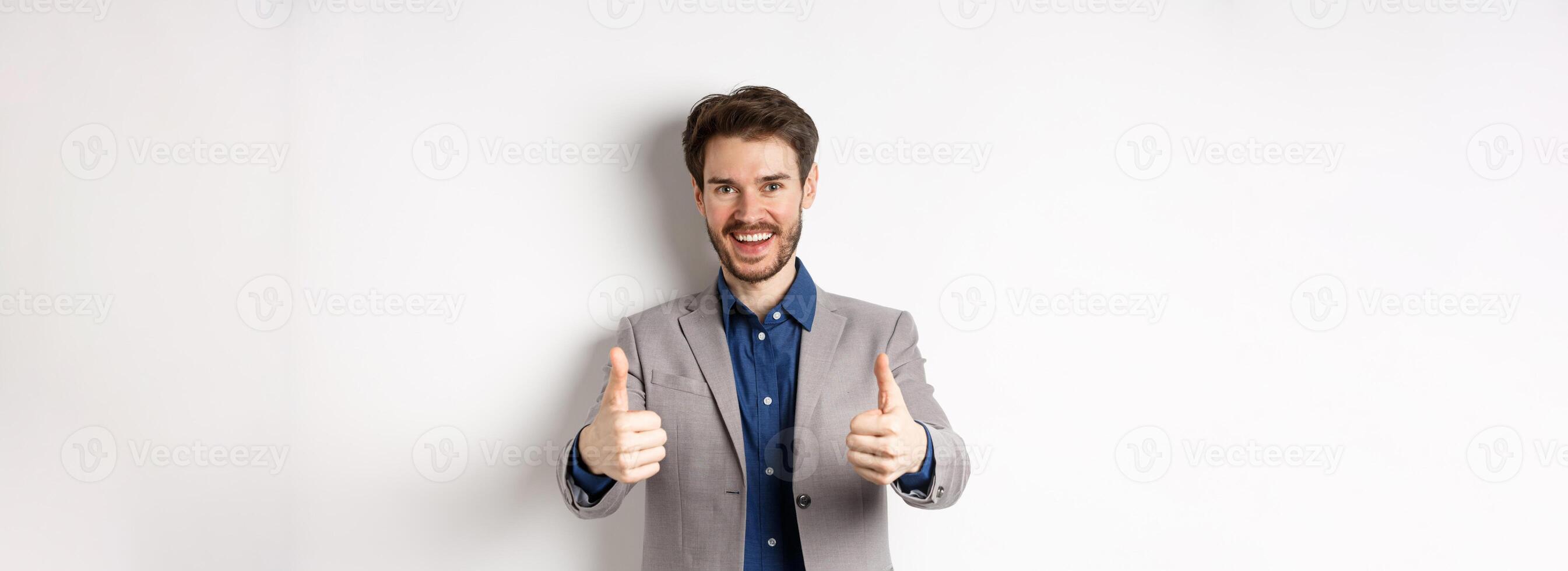Cheerful and positive young man in business suit showing thumbs up and smiling, like something good, praise good job, standing happy on white background photo