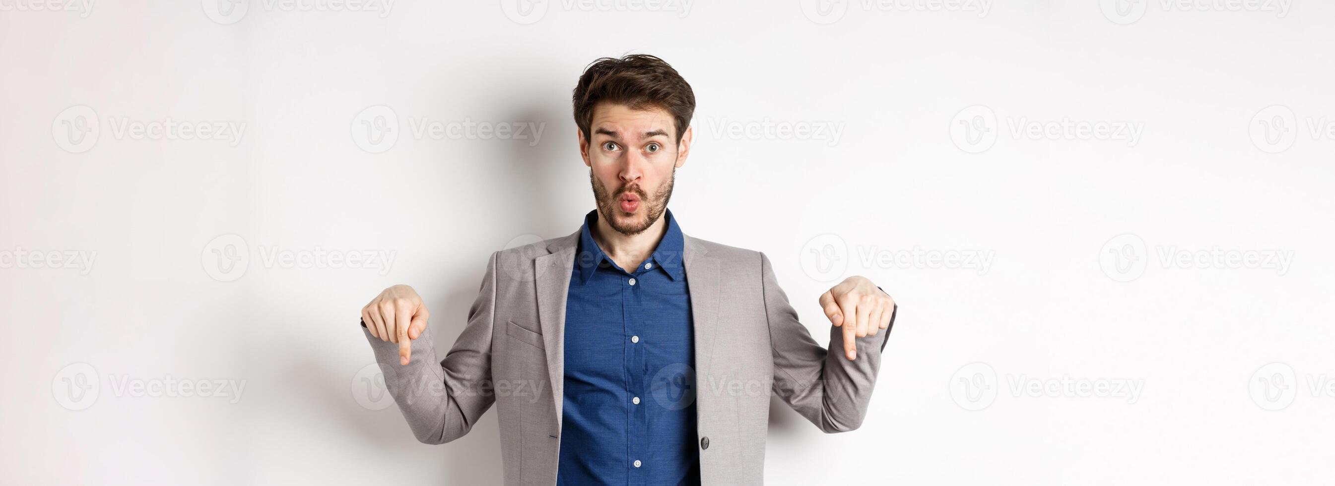 Wow look down. Excited business guy in suit pointing fingers bottom advertisement and look amazed, standing on white background photo