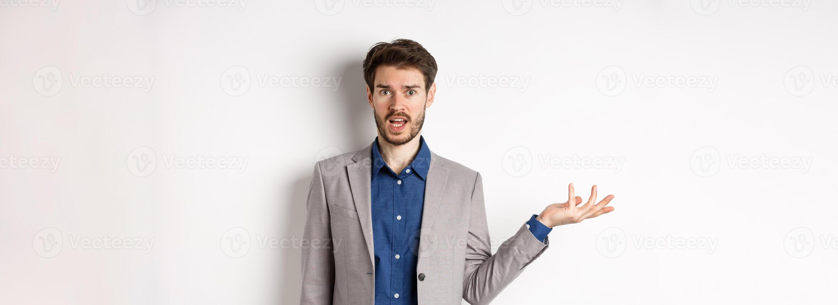 Whats wrong. Confused guy in suit cant understand, looking clueless with hand raised up, staring at camera questioned, standing on white background photo