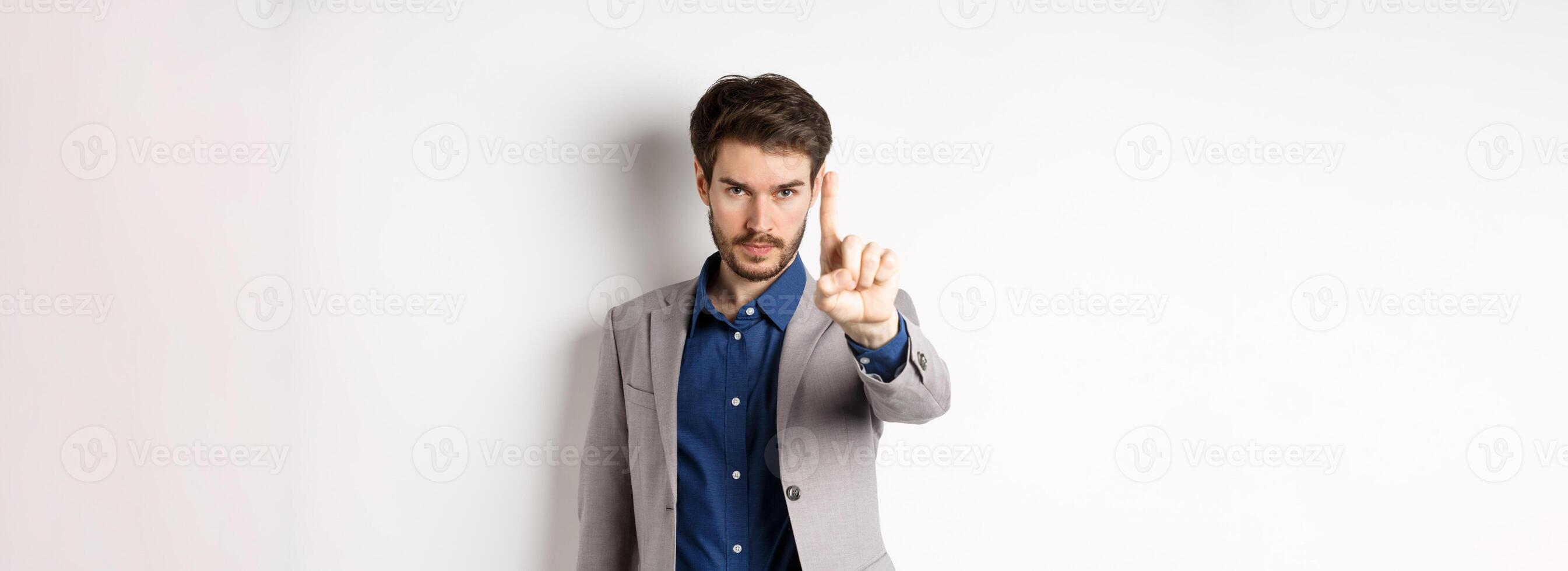 Listen to me. Serious male ceo manager showing one finger, scolding or telling to stop, tell no, hold right there sign, standing on white background in suit photo
