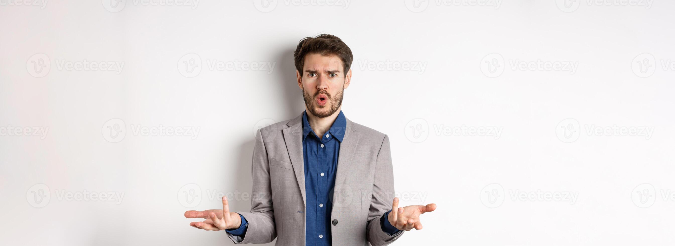 Confused and shocked businessman raising hands up and asking what happened, frowning disappointed, waiting for explanation, standing in suit on white background photo