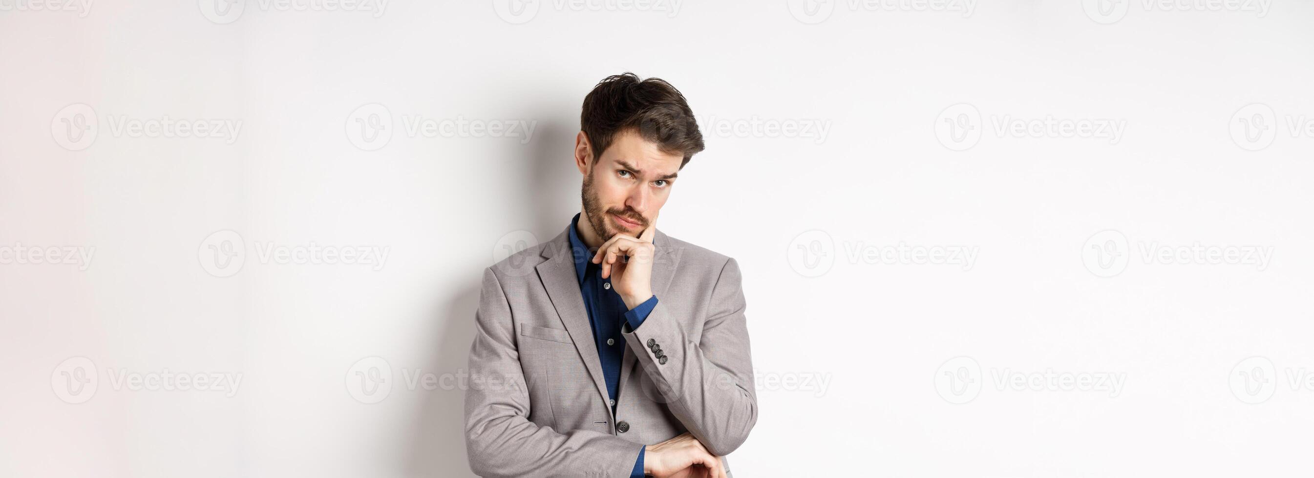 Hesitant businessman in suit making decision, looking pensive at camera with concerned face, having doubts, standing on white background photo