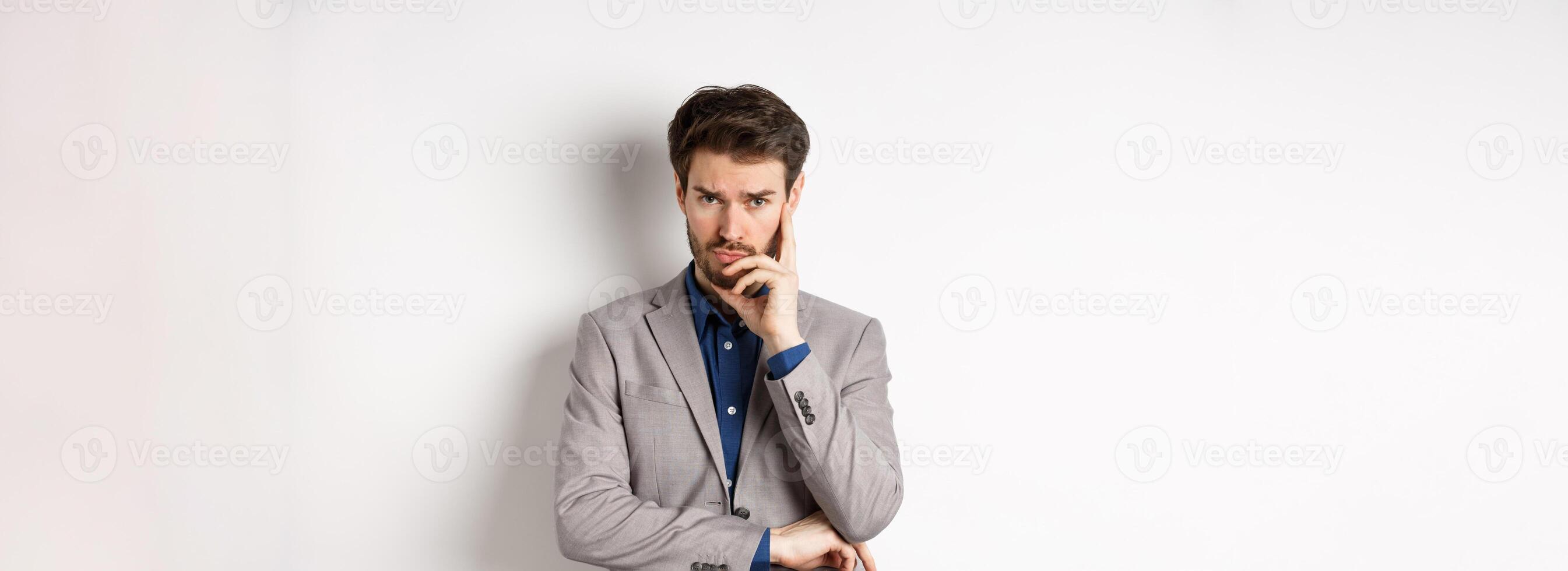 Troubled frowning business man in suit looking at camera pensive, thinking or making decision, standing on white background photo