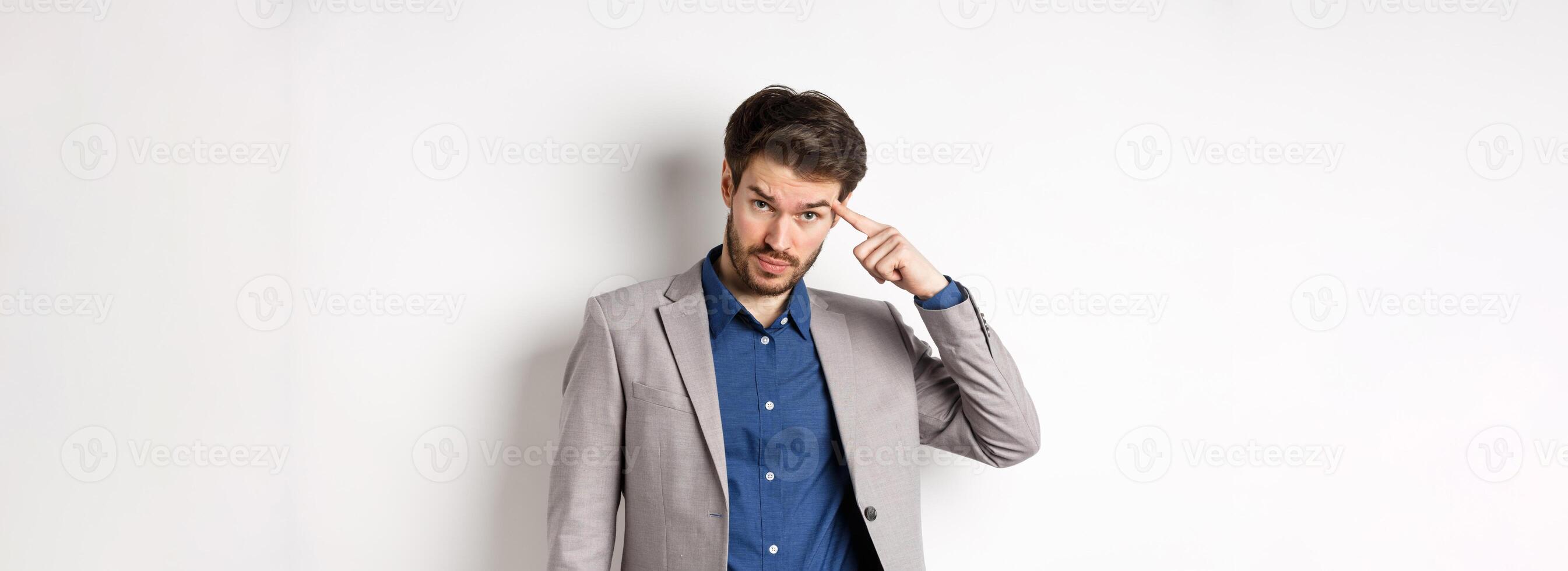 Annoyed male employer scolding personal, pointing finger at head, are you stupid out of your mind gesture, stare at camera from under forehead, standing in suit on white background photo