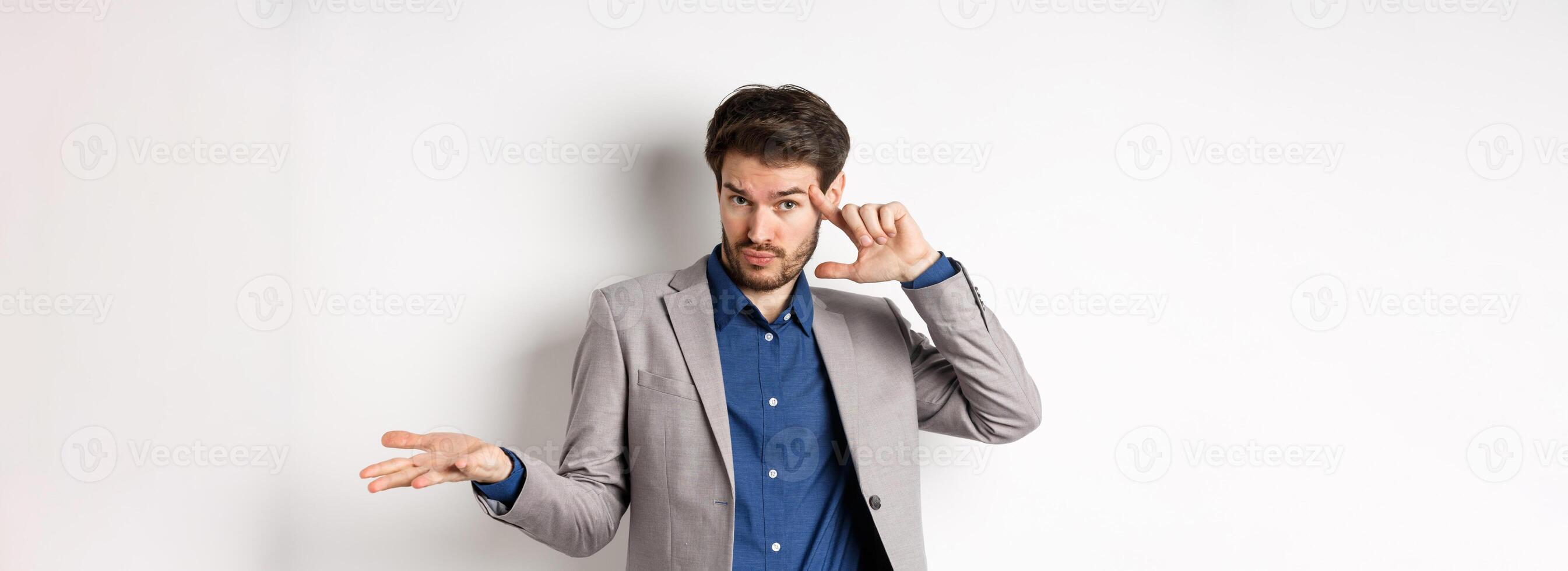 Are you crazy. Young man in suit pointing finger at head to scold strange or stupid mistake, shrugging shoulders confused, standing on white background photo