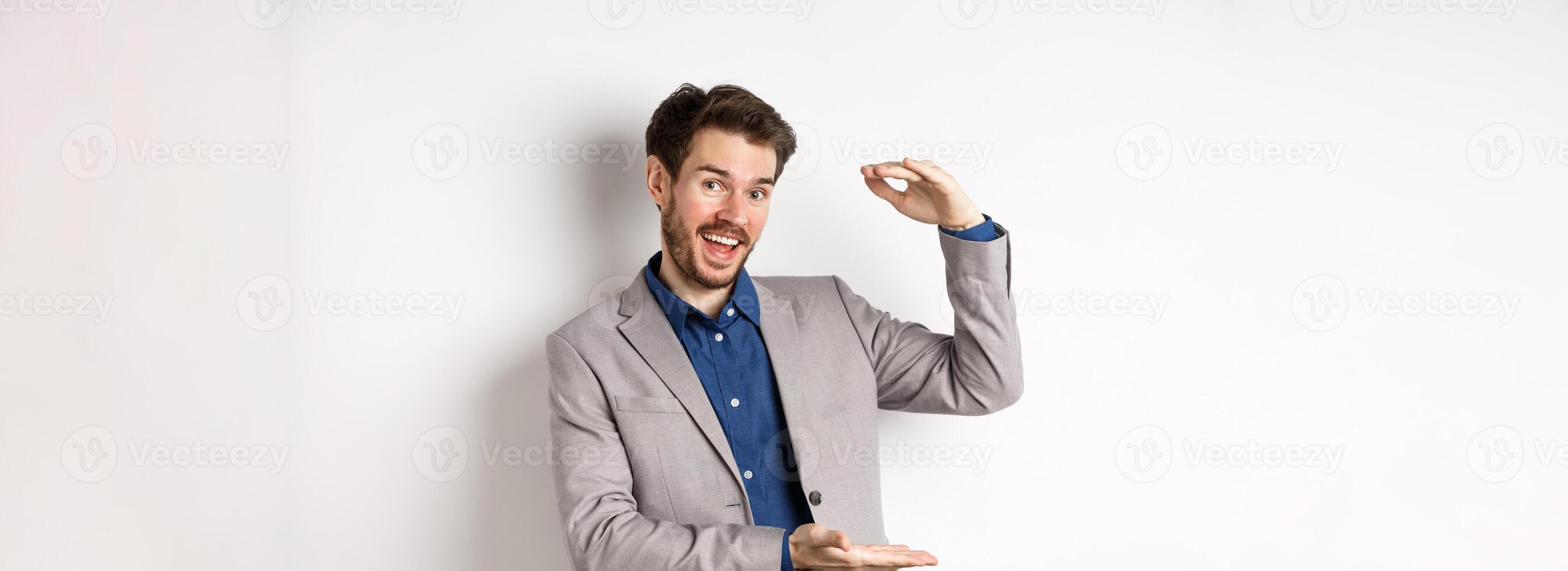 Handsome salesman in suit showing big size, large income, smiling at camera enthusiastic, standing on white background photo