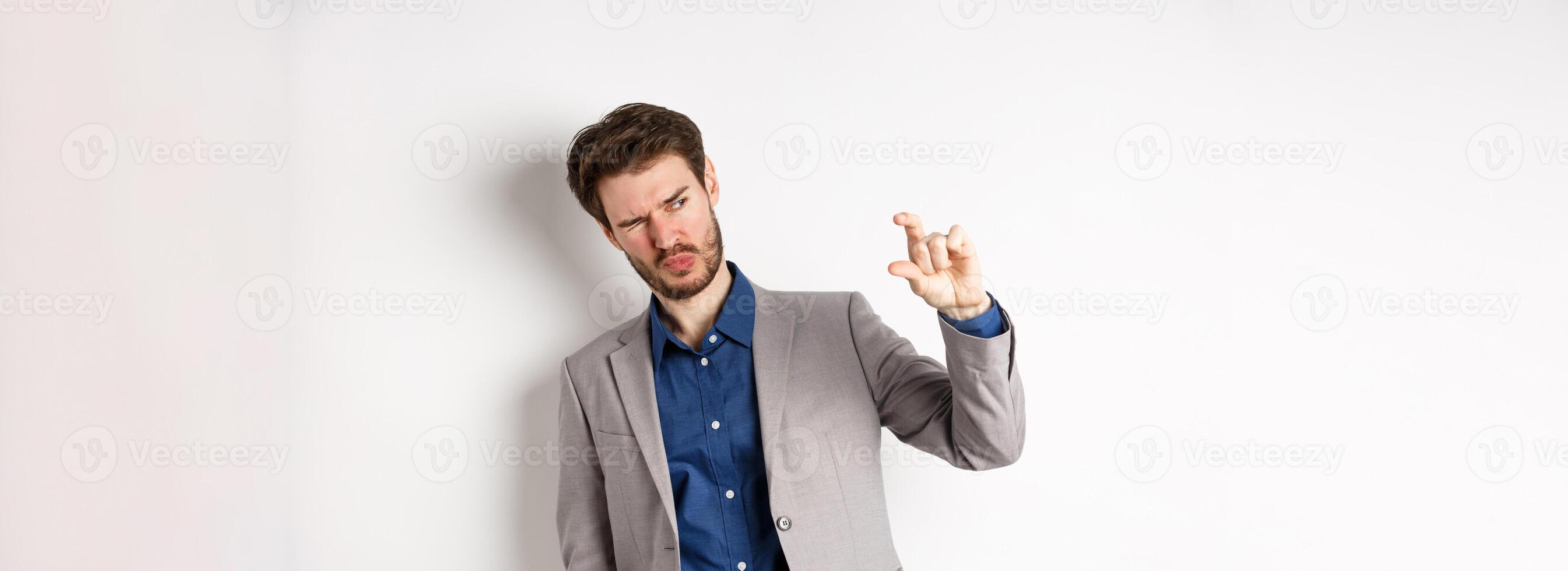 Businessman trying to see something tiny, showing small thing size with fingers, standing in suit on white background photo