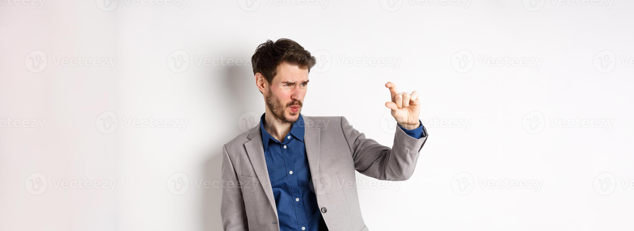 Businessman trying to see something small, showing little thing size with fingers, standing in suit on white background photo