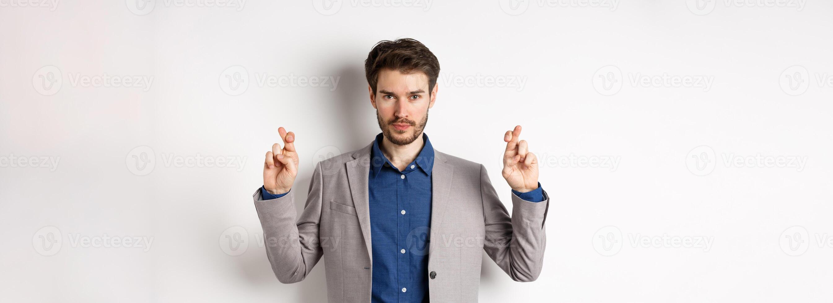 Hopeful man in suit cross fingers for good luck and looking confident in win, feeling determined, standing on white background photo