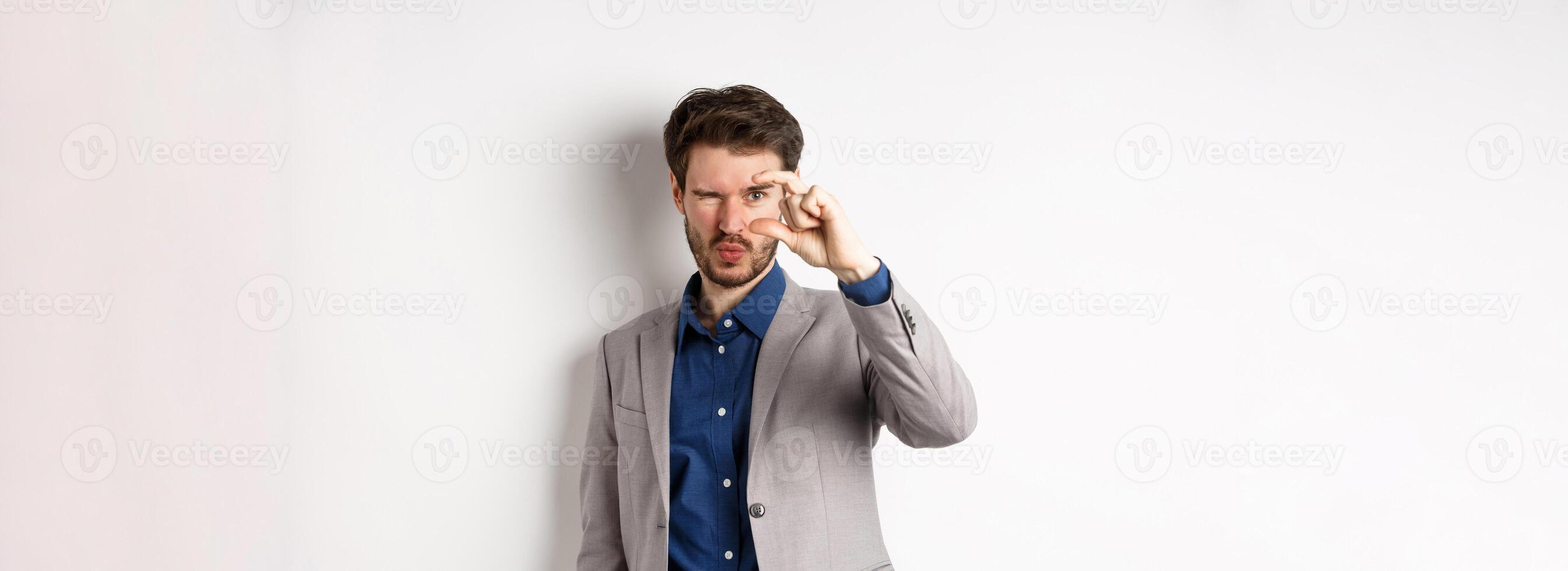 Businessman trying to see something tiny, showing small thing size with fingers, standing in suit on white background photo