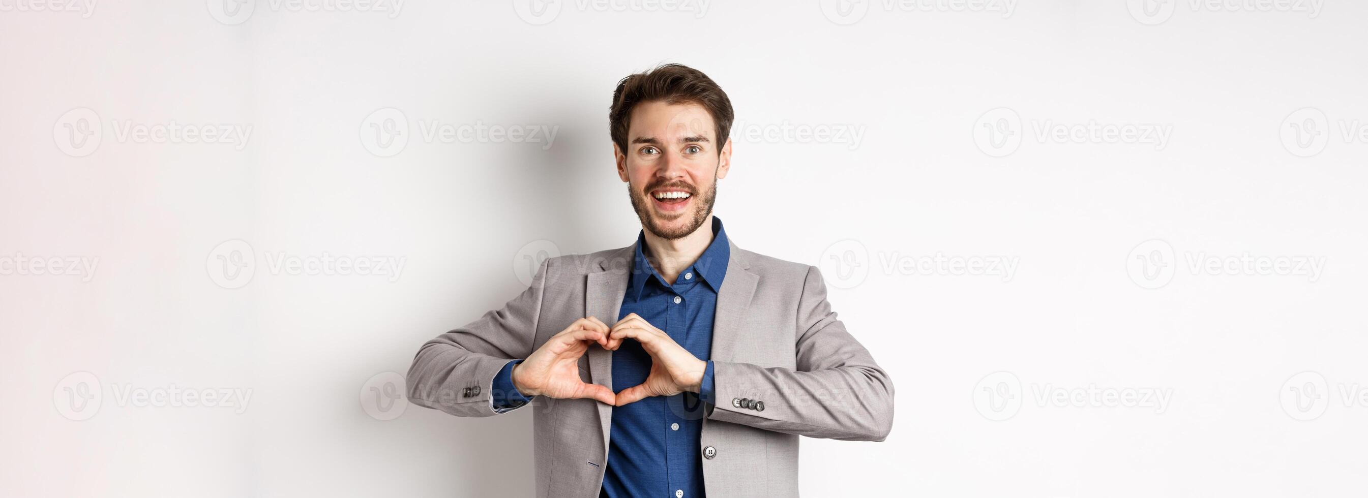 Passionate and romantic man in suit falling in love, showing heart sign and smiling at lover, standing on white background photo