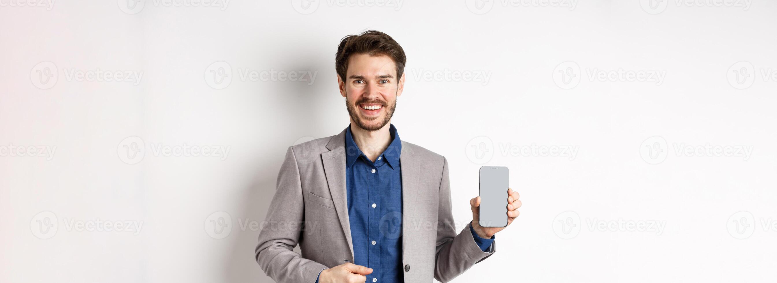 Handsome smiling salesman in suit showing empty smartphone screen, demonstrate an app, standing on white background photo