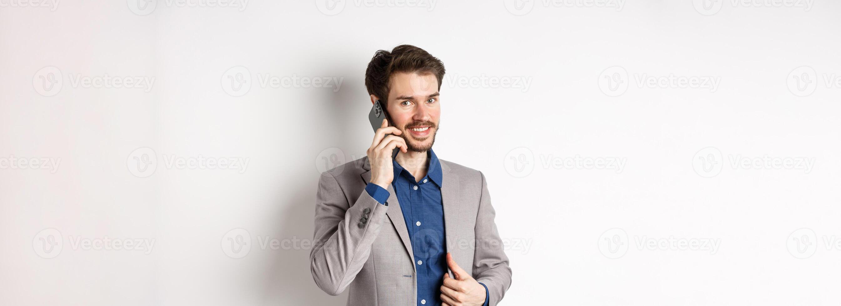 Successful businessman fixing his suit and talking on mobile phone, smiling at camera, standing on white background photo