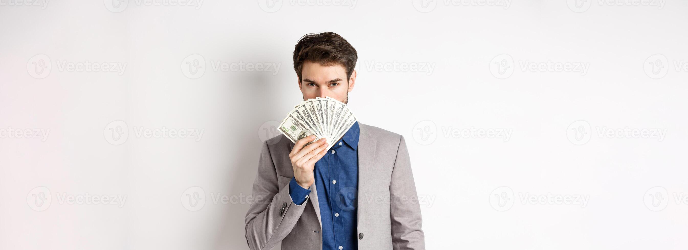 Rich businessman in suit showing dollar bills, hiding face behind money with sensual gaze at camera, white background photo