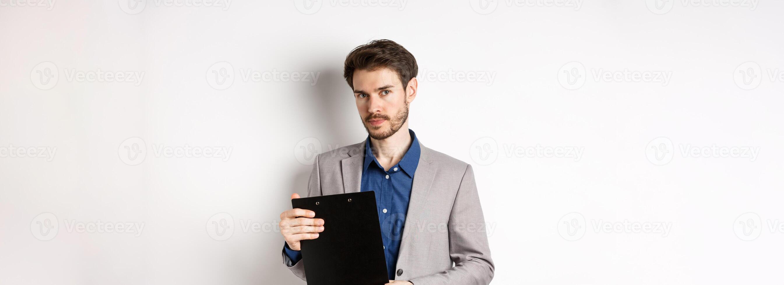 Young male ceo manager holding clipboard with documents and looking at camera, inspecting office, standing on white background photo