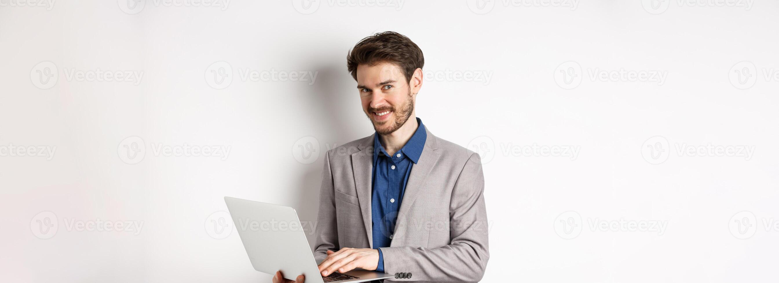 Successful smiling businessman working on laptop and looking happy at camera, standing in grey suit on white background photo