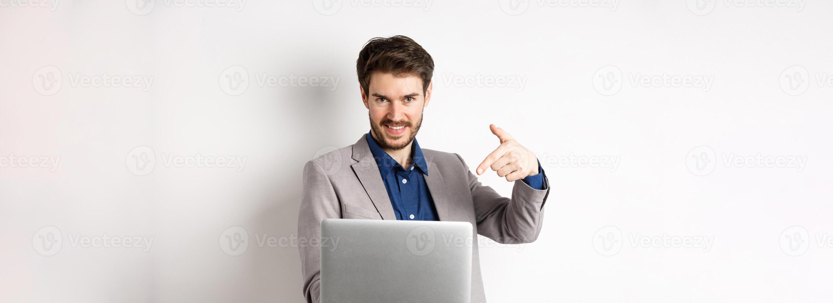 Confident young male entrepreneur working online with laptop, pointing at computer and smiling, standing on white background photo