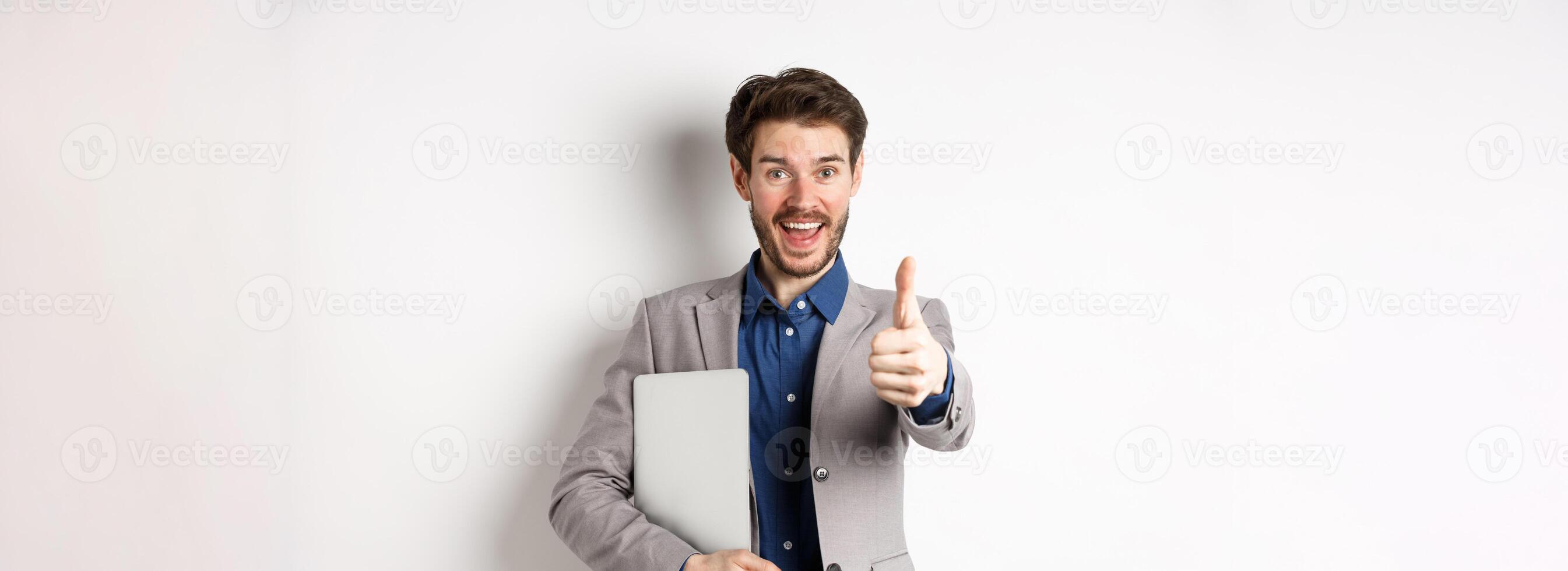 Good job. Happy smiling business man in suit holding laptop, showing thumb up to praise you, nice work gesture, standing on white background photo