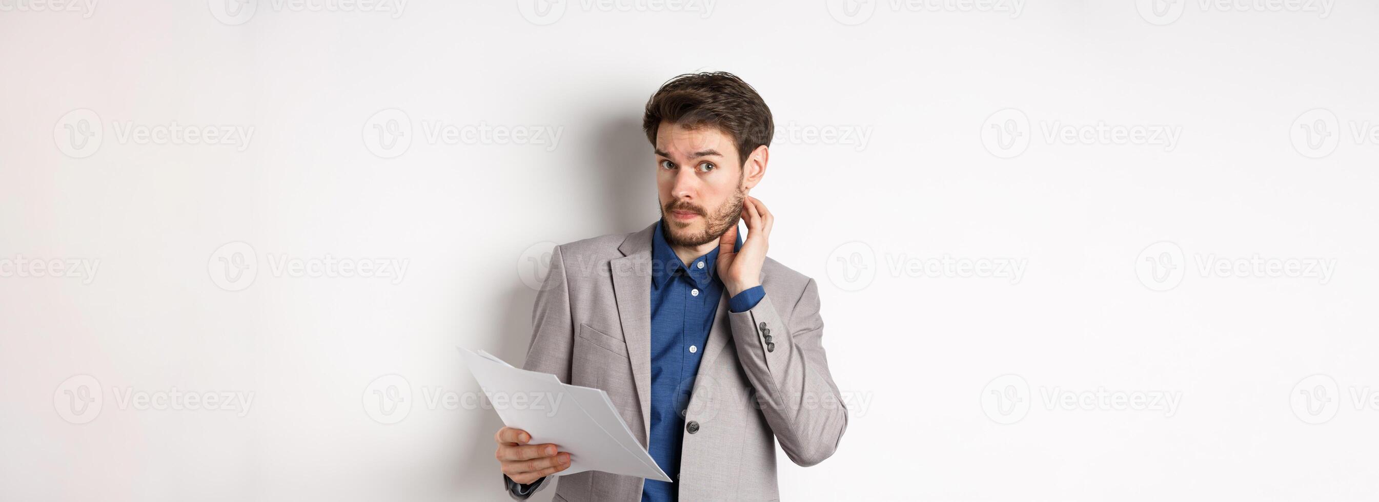 Confused office worker in suit scratching beard and look clueless, cant understand document, holding paper with indecisive face, white background photo