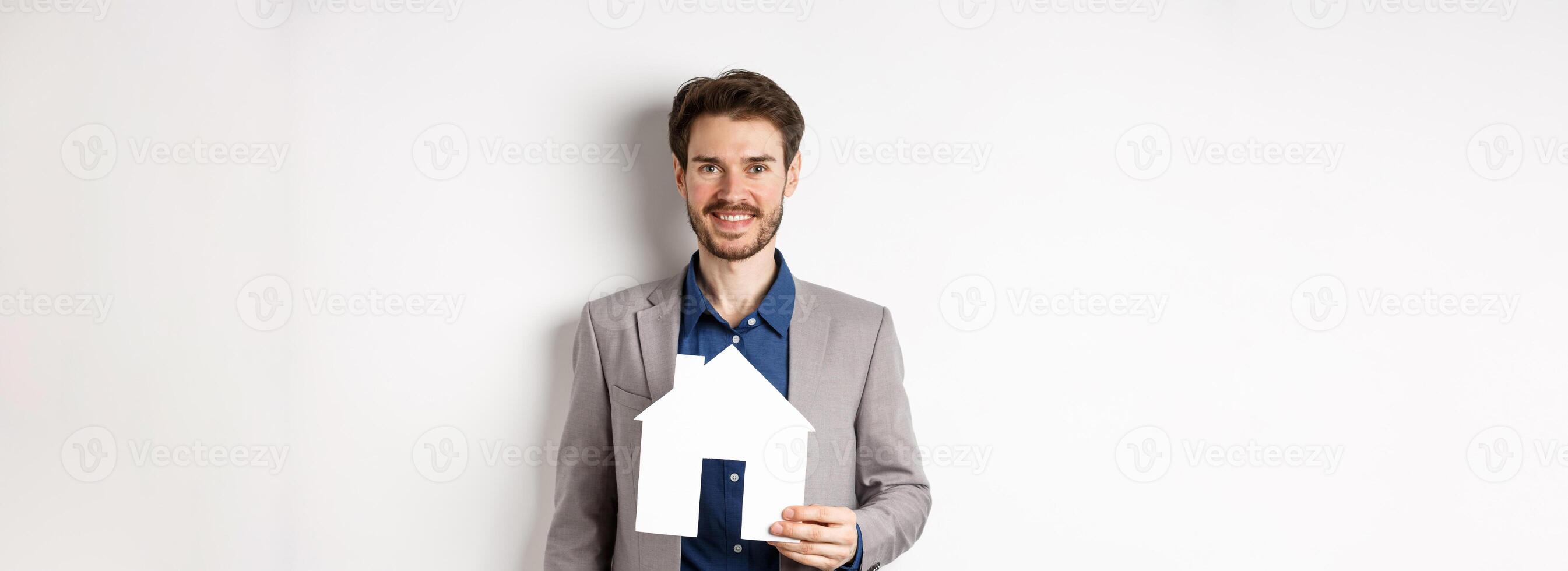 Real estate. Handsome young businessman buying property, holding paper house cutout and smiling, agency advertisement, white background photo