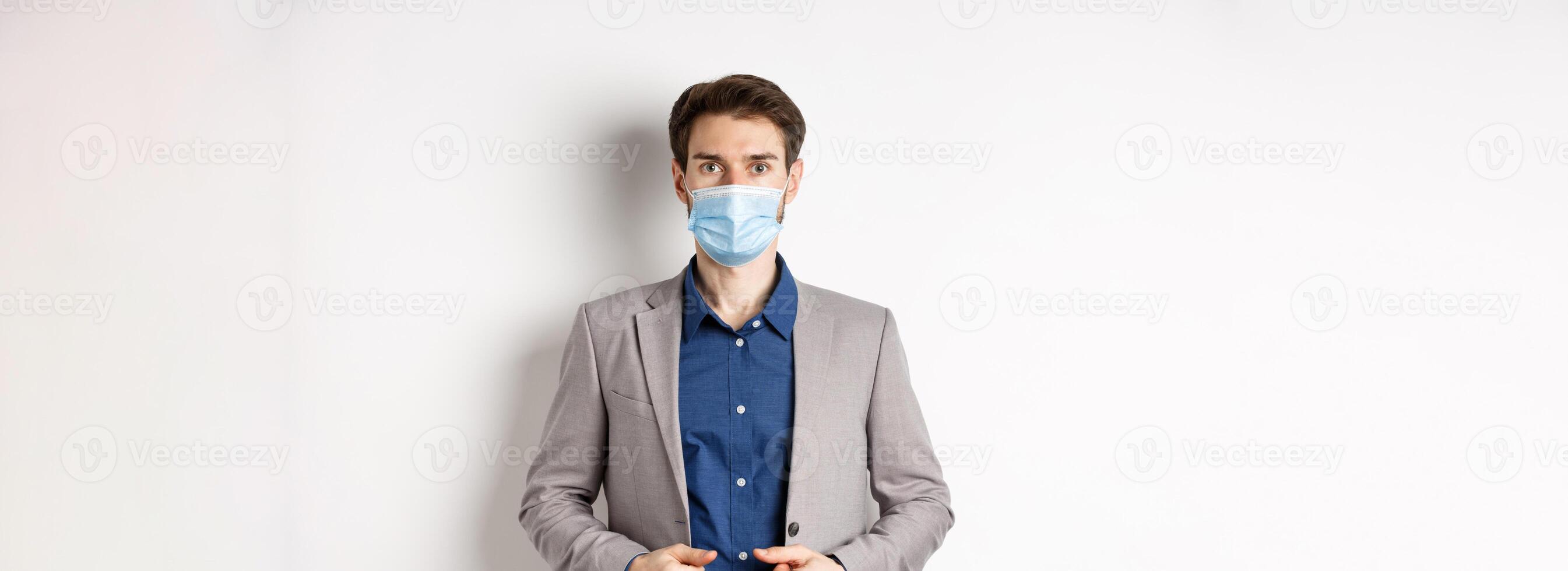 Covid-19 and office workers concept. Confident businessman in medical mask and suit looking at camera, standing on white background photo