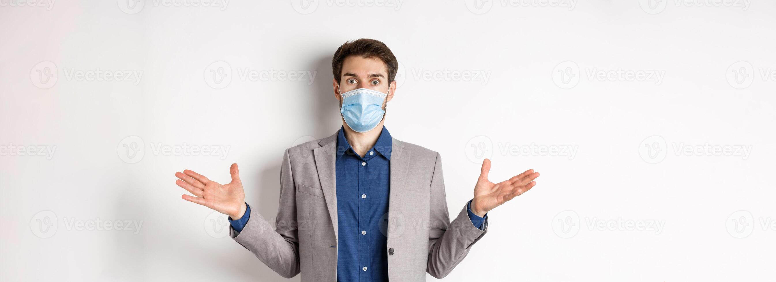 Health, pandemic and business concept. Confused businessman in medical mask and suit spread hands sideways and look puzzled, cant understand, white background photo