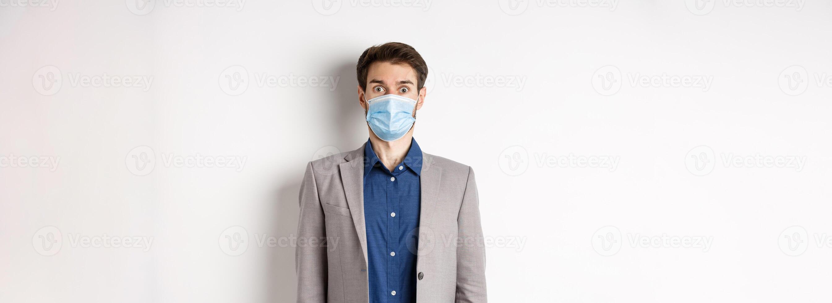 Health, pandemic and business concept. Surprised guy in suit and medical mask raising eyebrows, stare shocked at camera, white background photo