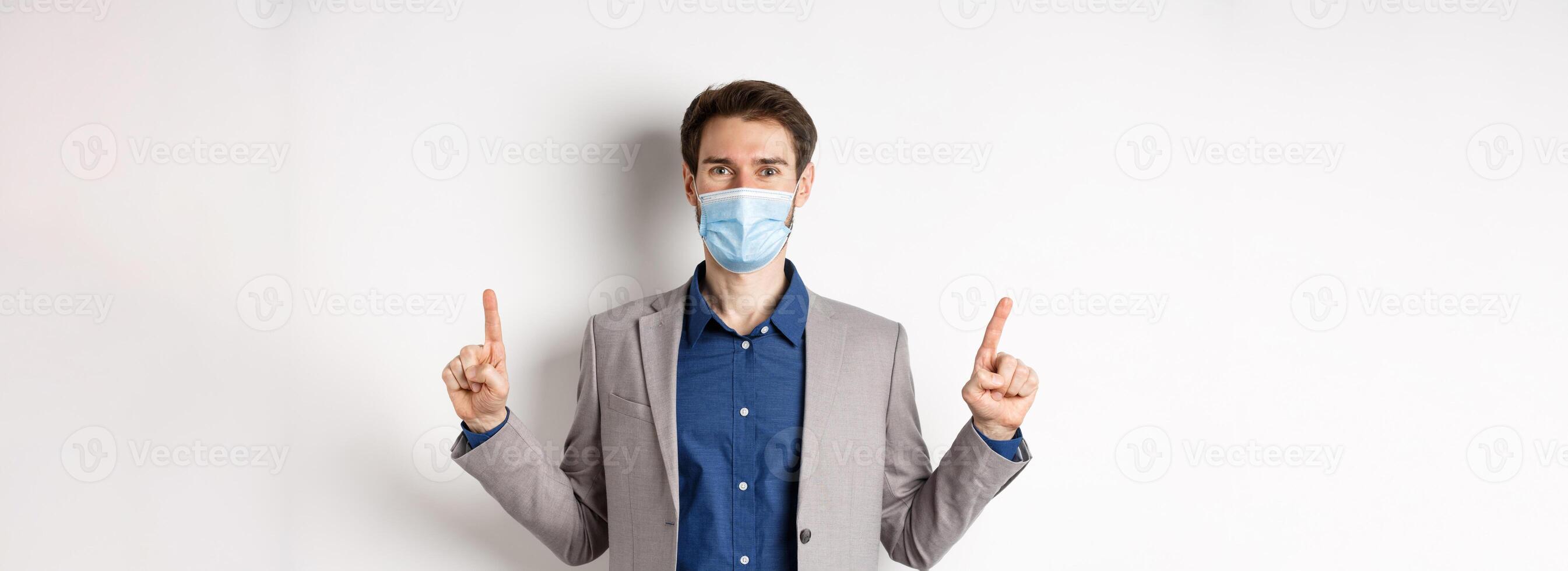 Covid-19, pandemic and business concept. Happy businessman in medical mask and suit pointing fingers up, standing on white background photo