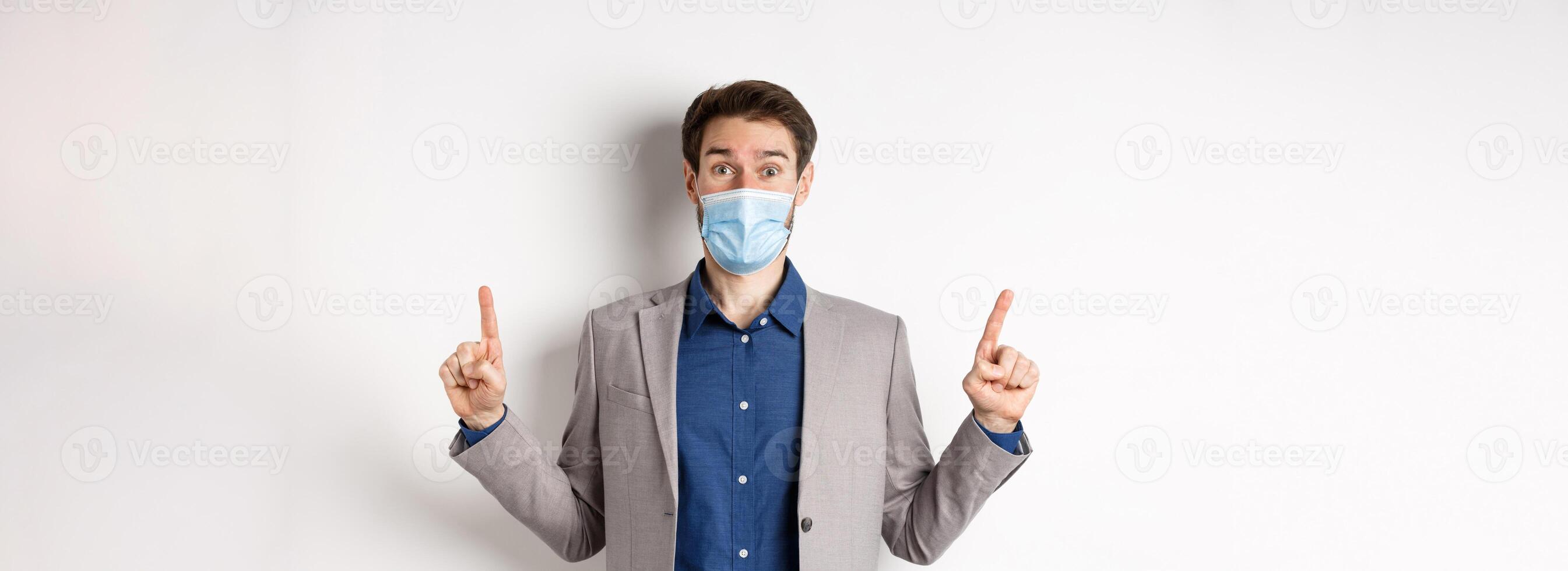 Covid-19, pandemic and business concept. Excited businessman in sterile medical mask and suit pointing fingers up, white background photo
