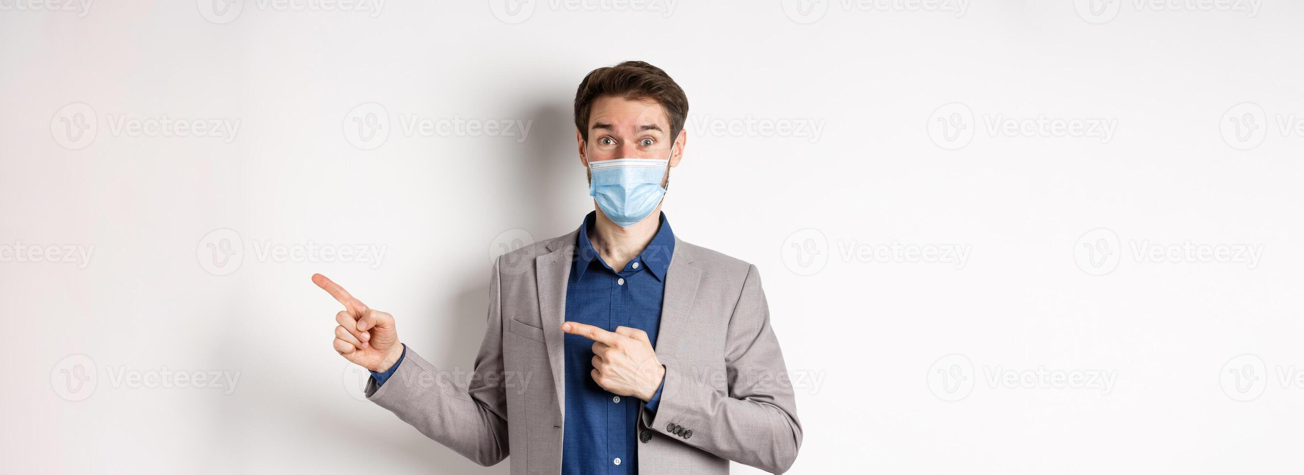 Covid-19, pandemic and business concept. Excited male entrepreneur in medical mask and suit pointing fingers right and looking curious, white background photo