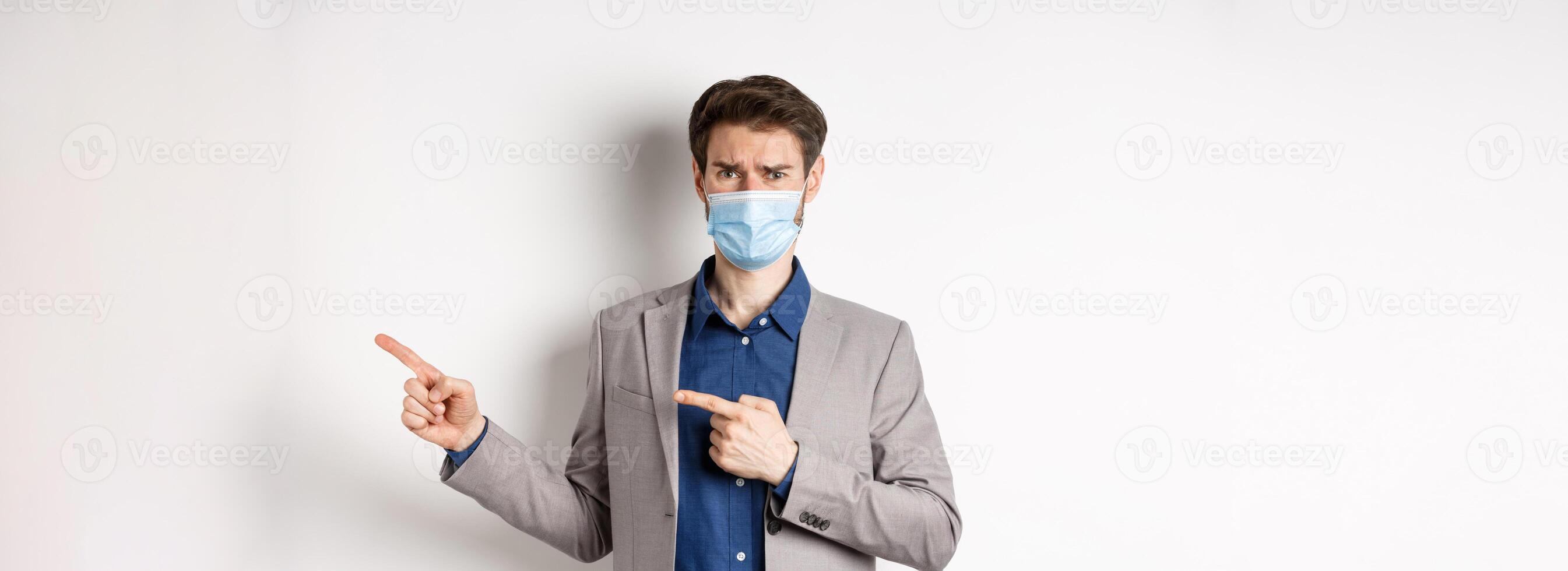 Covid-19, pandemic and business concept. Disappointed and reluctant man in medical mask frowning, pointing fingers right at something disgusting or ugly, white background photo
