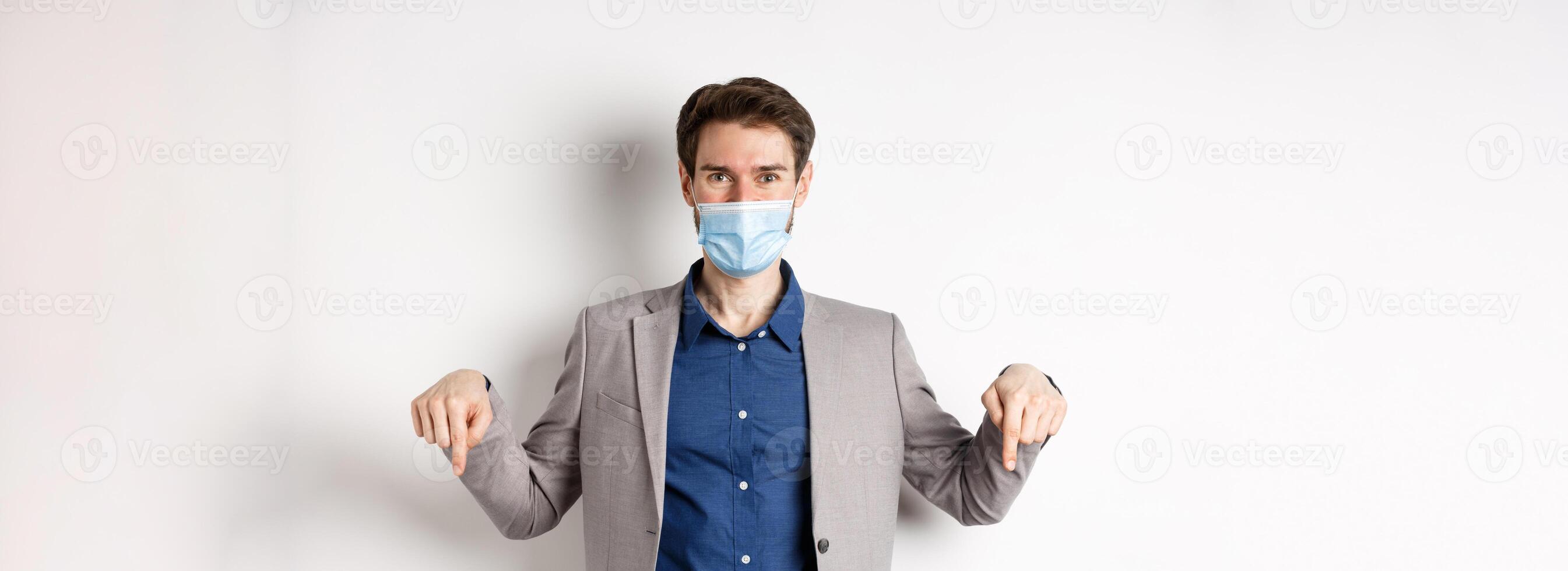 Covid-19, pandemic and business concept. Handsome businessman in medical mask and suit, pointing fingers down and showing company logo, white background photo