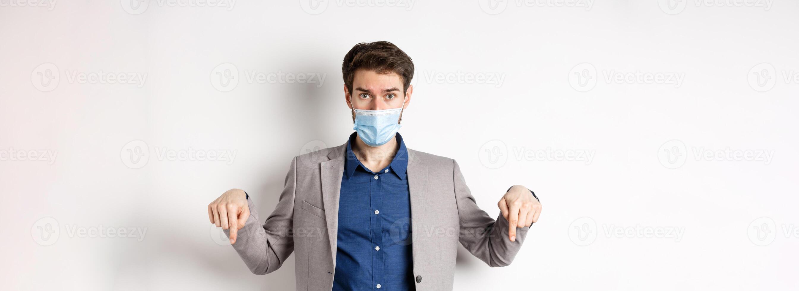 Covid-19, pandemic and business concept. Hesitant man in suit and medical mask asking opinion, pointing fingers down and look at camera, white background photo