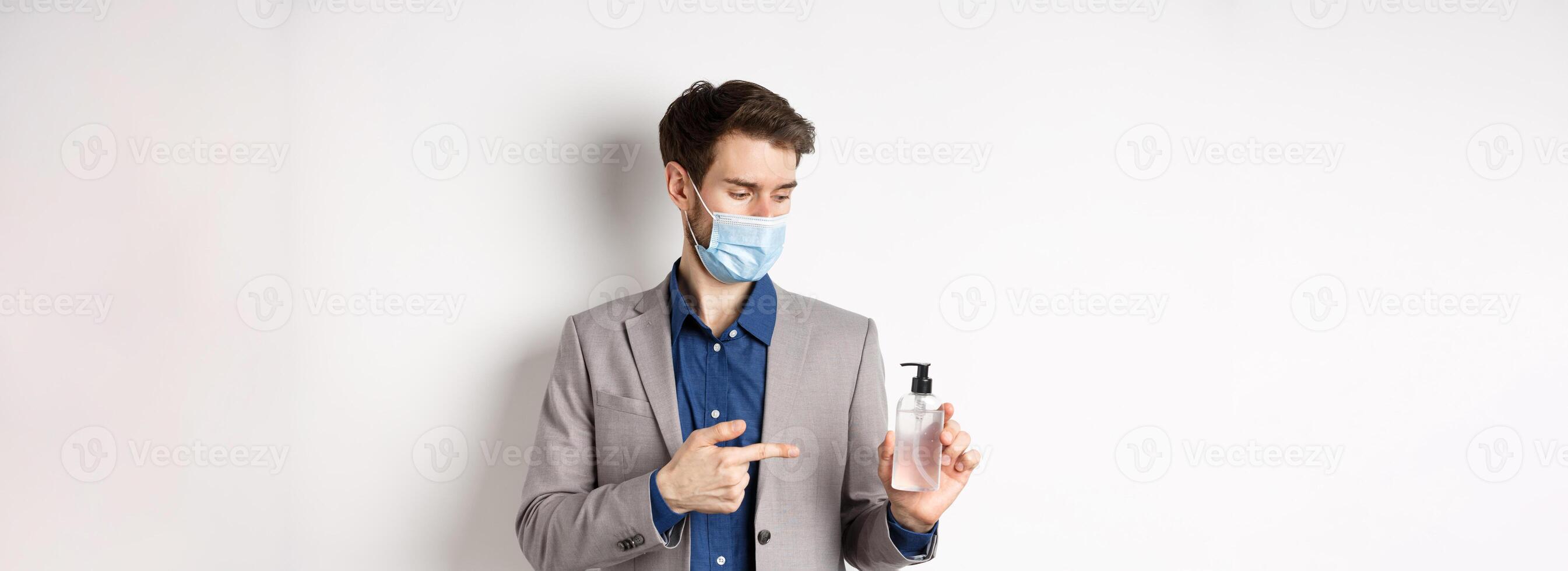 Covid-19, pandemic and business concept. Office worker in medical mask pointing at bottle of hand sanitizer, using antiseptic at work, white background photo
