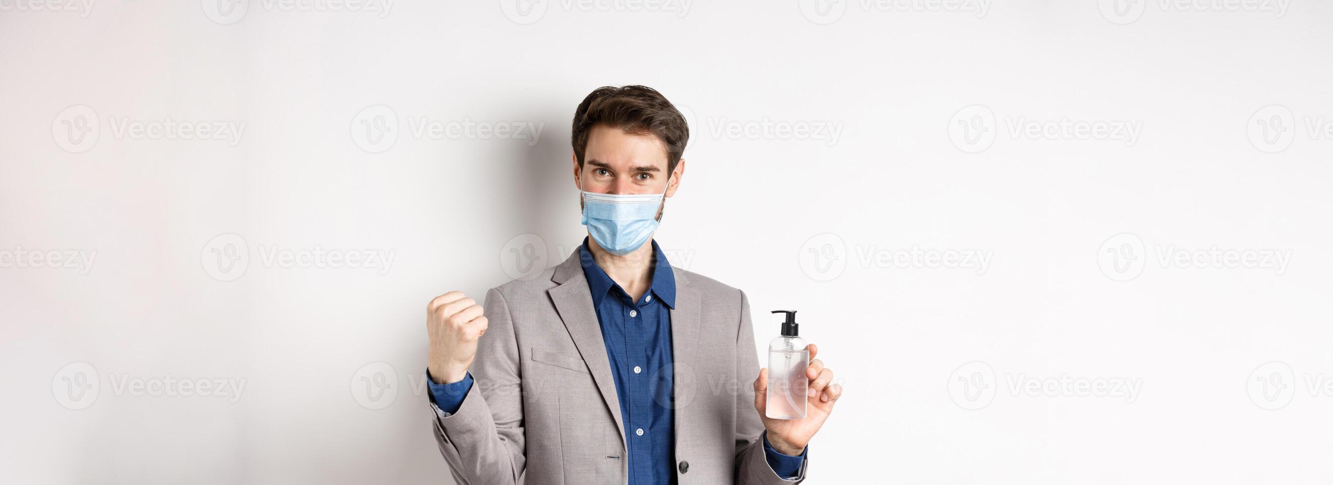 Covid-19, pandemic and business concept. Cheerful office guy in medical mask and suit, looking motivated, showing bottle of antiseptic, white background photo