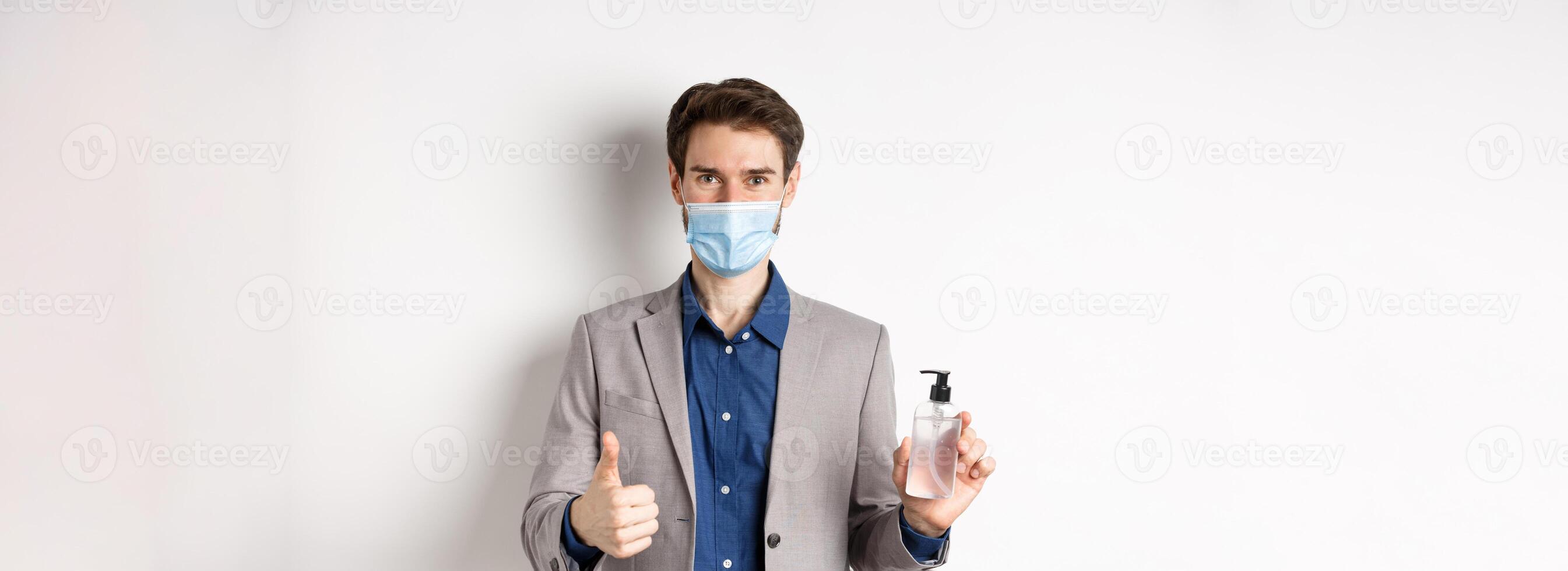 Covid-19, pandemic and business concept. Businessman in office suit and medical mask showing bottle of hand sanitizer and thumb up, recommend use antiseptic at work photo